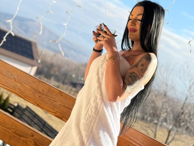 ArianaTattoos with long black hair, white towel dress, holding a glass, tattoos on right arm, standing on wooden porch, sunlight, winter, mountains in background.