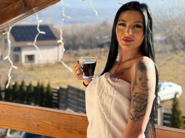 ArianaTattoos with long black hair, pink lipstick, and a tattooed arm, holding a beer glass, wearing a white towel, standing on a wooden balcony.