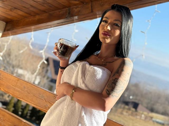 ArianaTattoos with long black hair, holding a beer mug, wearing a white towel, tattoos on arm, sunny outdoor wooden porch, looking at camera with red lipstick.