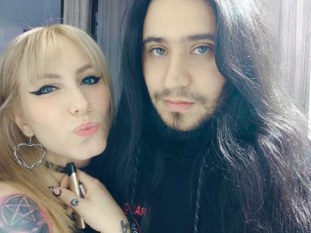 EmilyAndChrisHot has blonde hair, blue eyeliner, heart earrings, nose ring, lip piercing, holding a lipstick, pouty lips. ChrisHot has black hair, beard, blue eyes, black shirt.