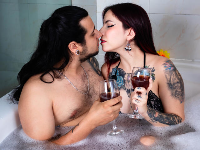 EmilyAndChrisHot with tattoos, in a bubble-filled bathtub, kissing while holding glasses of red wine. Chris has long black hair, Emily has red hair, both are topless.