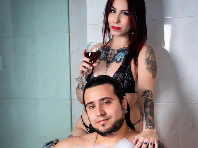 EmilyAndChrisHot with tattoos, black lace bra, red wine, standing behind ChrisHot, who's shirtless with tattoos, in a bathroom. ChrisHot has a beard and black hair.