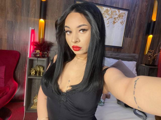 MadyLux hat schwarzes Haar, roten Lippenstift und trägt ein schwarzes Tanktop, das ihre Brüste zeigt. Sie hat Tattoos an ihrem Arm und trägt eine Halskette. Sie macht ein Selfie..