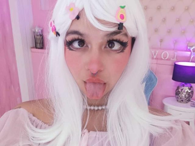 LaraAhB a les cheveux blancs, de grands yeux avec de longs cils, un anneau au nez, et elle tire la langue. Elle porte un haut rose à une épaule et un collier de perles..