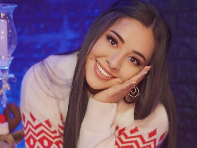 SantaRodrigues has long black hair, brown eyes, and a cute smile. She's wearing a white sweater with red zigzags, resting her cheek on her hand, looking cute.