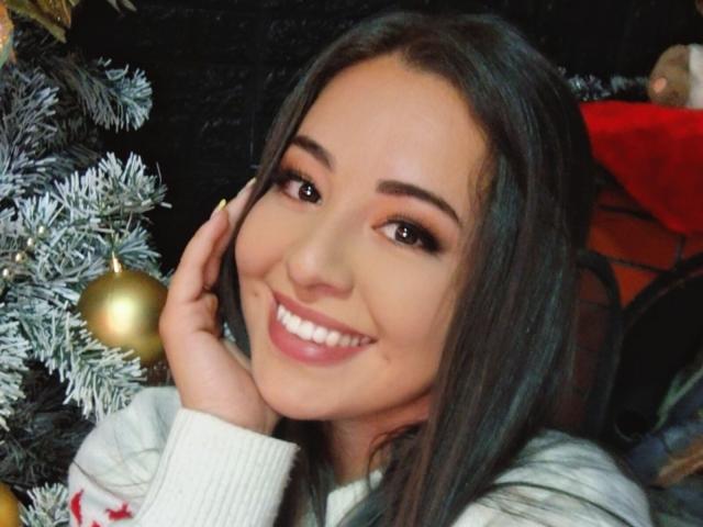 SantaRodrigues, com longos cabelos escuros, sorrindo com lábios rosados, olhos castanhos e um suéter branco, tocando a bochecha, ao lado de uma árvore de Natal coberta de neve com enfeites dourados..