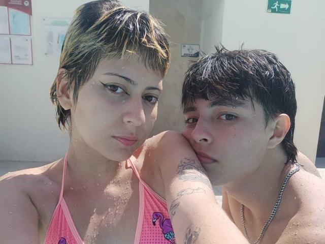 JamesAndRouse with short, wet hair, pink bikini top, small tits, tattoo on arm, leaning on JamesAndRouse with wet black hair, nose piercing, no shirt, in a sunlit bathroom.