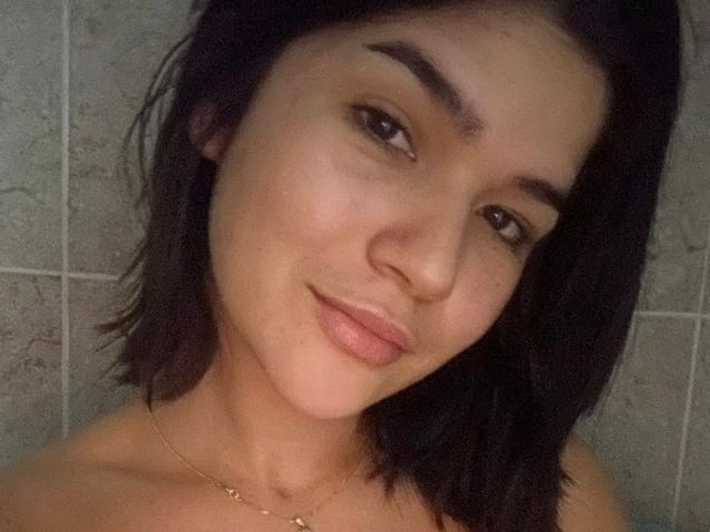ScarlletZhen has medium brown skin, black shoulder-length hair, and a subtle smile. She's wearing a gold necklace and has a slight nose tilt. Her tits are not visible.