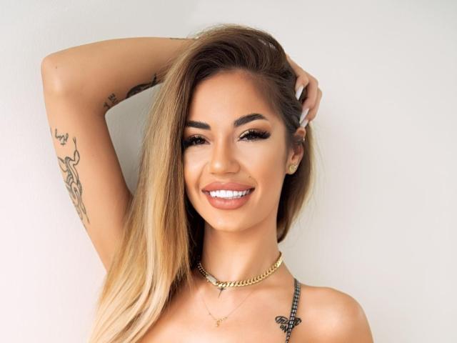 Yuukenzi has long blonde hair, a gold chain necklace, and a tattoo on her right arm, smiling with her arm raised. She has white teeth and full eyebrows.