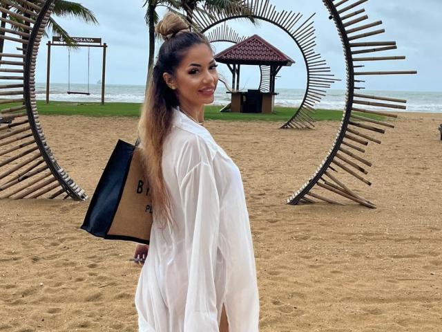 Yuukenzi in a wet white shirt, showing her tits, holding a black bag, standing on a beach with a wooden circle structure, palms in the background, ocean behind.