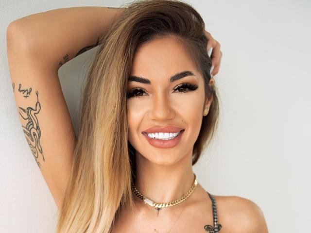 Yuukenzi has a cute smile with white teeth, long blonde hair, and a tattoo on her arm. She's wearing a gold necklace and has her arm raised.