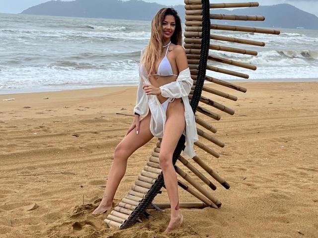 Yuukenzi with long blonde hair, white bikini, and sheer shirt, standing on sandy beach, leaning on wooden and black rope structure, smiling, showing her tits and ass.