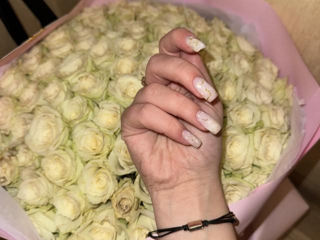 SugarrX's hand with pale, glittery nails and a black bracelet holding a pink bouquet of white roses.