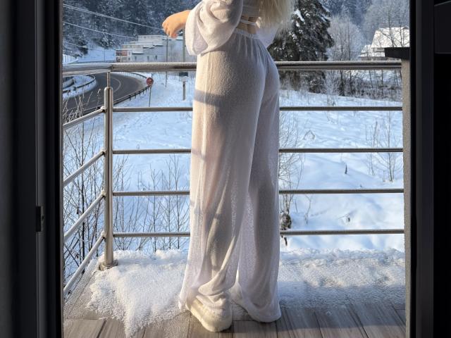 SugarrX in white, sheer, loose pants, showing her ass, standing on a snowy balcony, facing outside, with metal railing in front and wooden floor beneath.
