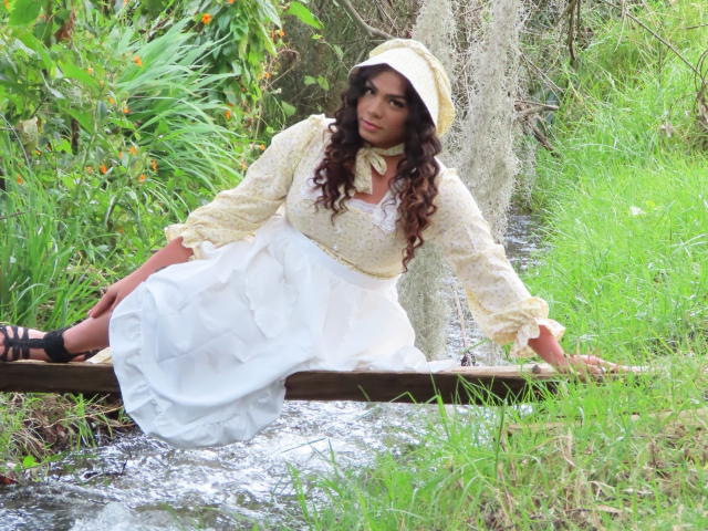 LiliithBuckland with curly brown hair, white dress, and yellow top, sitting on a wooden plank in a green, grassy, and water-filled area, wearing black sandals.