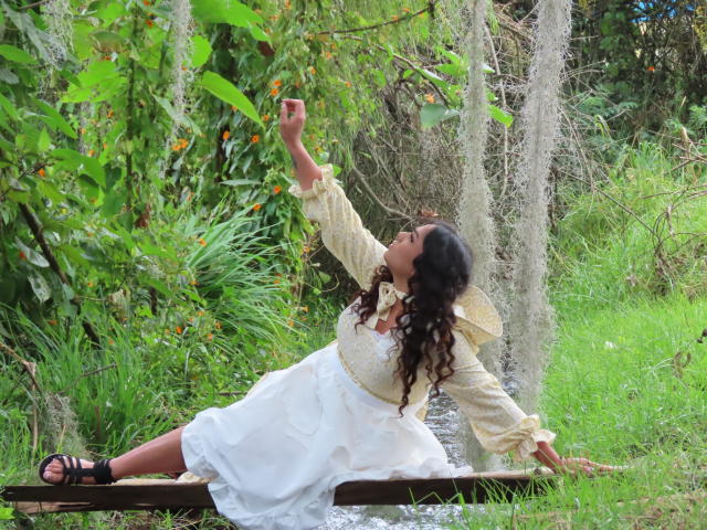 LiliithBuckland with curly black hair, white dress, and black sandals, leaning back on a wooden plank, arm raised, touching orange berries on a green plant.