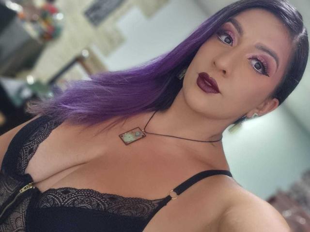 AndreaSharick with purple hair, black lace bra, deep red lipstick, and a necklace, showing her tits and a confident look. Blurred background.
