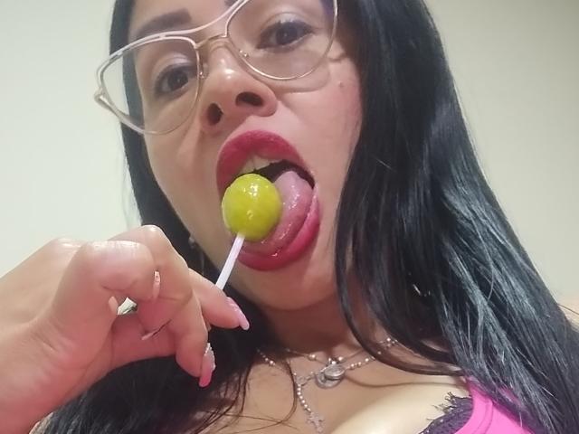 KinkyVane, with glasses, black hair, and pink lipstick, sucks a yellow lollipop, tongue out, hand holding stick, showing cleavage in pink lingerie.