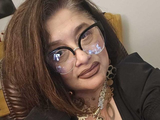 ClassybutNaughty, avec des cheveux bruns, des lunettes et un rouge à lèvres foncé, portant un haut noir et un collier en or, avec un léger sourire et une expression détendue, un clin d'œil..