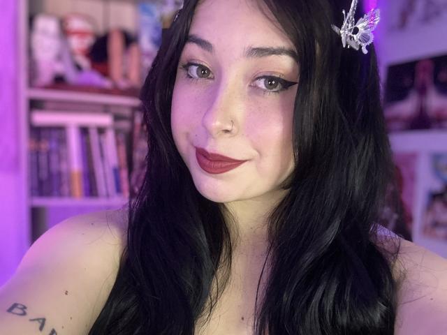 LuhannaGrise with black hair, butterfly hair clip, dark red lipstick, eyeliner, nose ring, and tattoo on shoulder, smiling at camera, purple and pink lighting, showing bare shoulders.