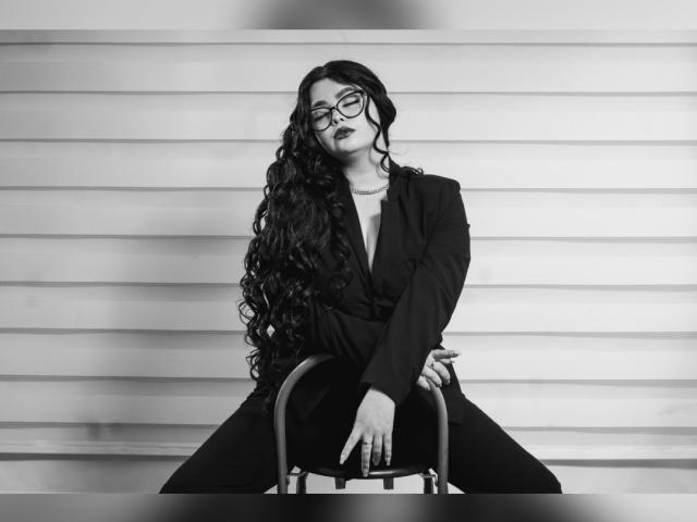 Fotografía en blanco y negro de BrithneyLee con cabello largo y rizado, usando gafas, lápiz labial oscuro, un blazer negro y pantalones, sentada en una silla, con las manos sobre el asiento y las piernas separadas..
