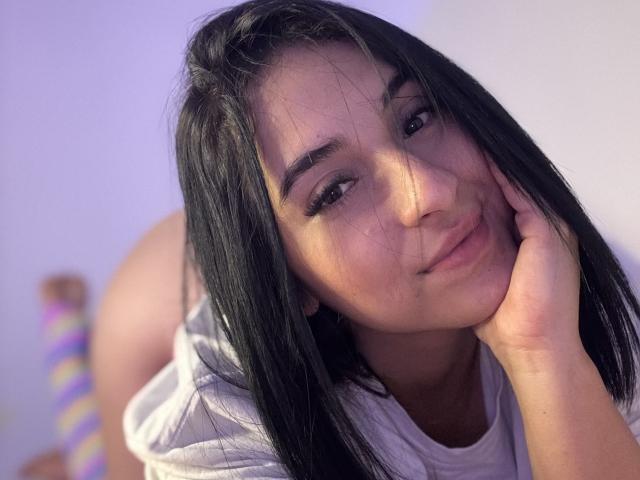 Emma Waltter, con i suoi lunghi capelli neri, il viso carino e il sorriso dolce, appoggia la guancia sulla mano, indossa una camicia bianca, e sullo sfondo si intravedono il suo sedere e un calzino colorato a righe..