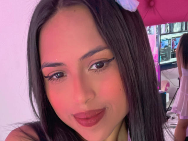 MyaaTyler with dark straight hair, pinkish-red nose, and thick black eyeliner, smiling with red lipstick, wearing a pink hair clip, in a pink-lit selfie.