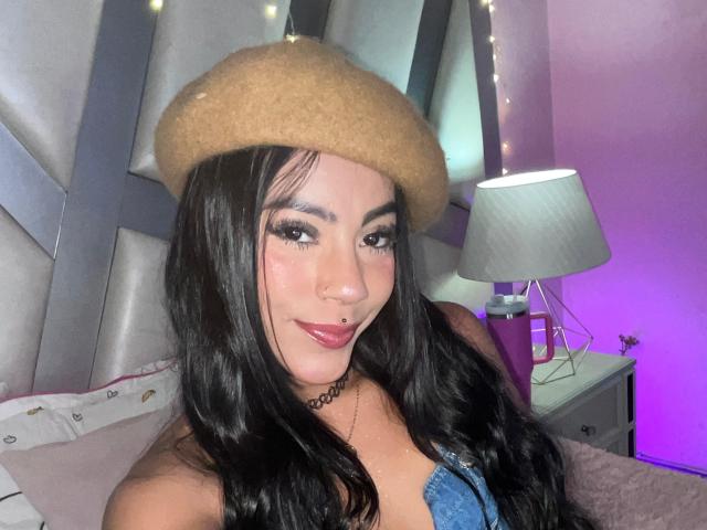 Amberv with long black hair, wearing a beige beret, has tits visible in a blue top, nose ring, and choker, smiling with pink lipstick in a purple-lit room.