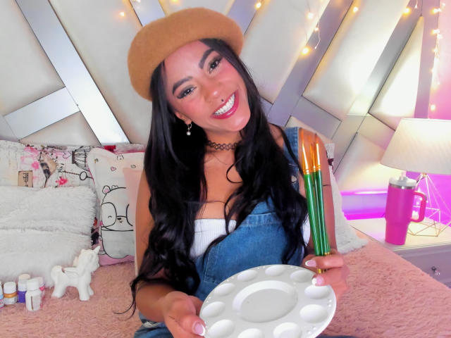 Amberv with long black hair, tan beret, denim overalls, white top, choker, holding white plate and colorful pens, smiling, pink lips, cute, sexy, bed with pink blanket, white pillow.