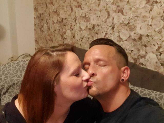 AgabeyCouple with red hair kissing AgabeyCouple with shaved head and ear gauges, both wearing black shirts, eyes closed, nose touching, soft expression, in a cozy indoor setting.