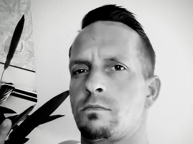 Black and white selfie of AgabeyCouple with short, dark, slicked-back hair, slight beard, and serious expression, holding a plant on the left side of the frame.