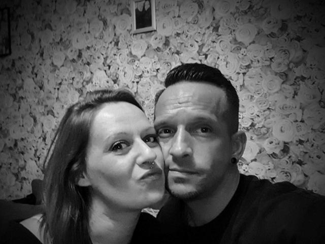 Black-and-white photo of AgabeyCouple, showing a woman with nose ring and a man with short, slicked-back hair, both with ear piercings, posing closely.