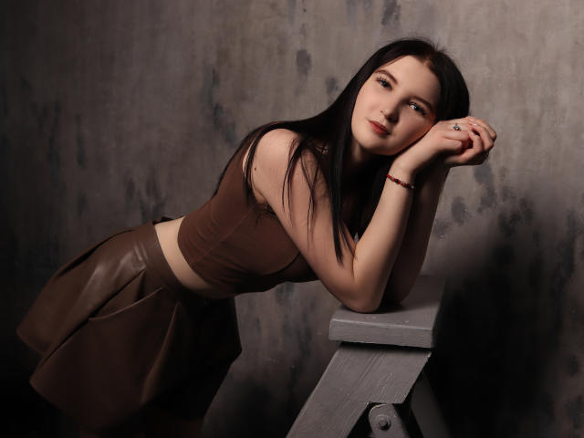 AngelaJones with long black hair, brown crop top, and leather skirt, leaning on a wooden stool, looking to the side with a slight smile, hands near her face.