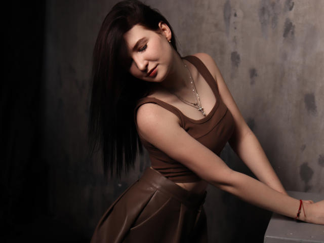 AngelaJones with brown hair, leaning forward, wearing a brown tank top and leather skirt, showing her small tits, red lipstick, necklace, and red bracelet, in a dark setting.