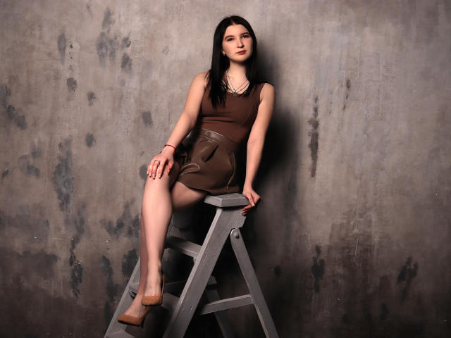 AngelaJones with dark brown dress, sitting on gray ladder, legs apart, showing her pussy, black hair, pale skin, brown shoes, and a necklace, against a textured wall.