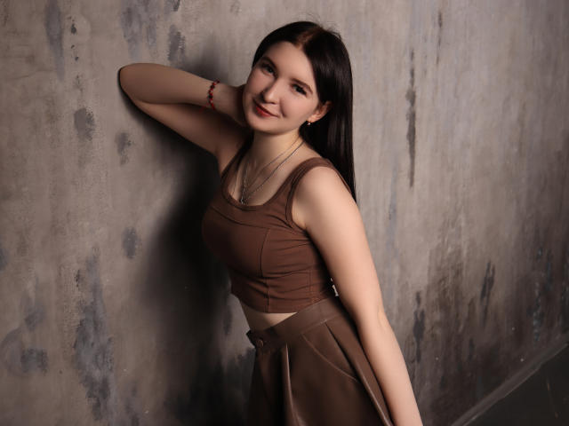 AngelaJones with long dark hair, brown crop top, and brown skirt, smiling, arm raised, touching wall, red bracelet, earrings, and a flat ass. Soft lighting, casual pose.