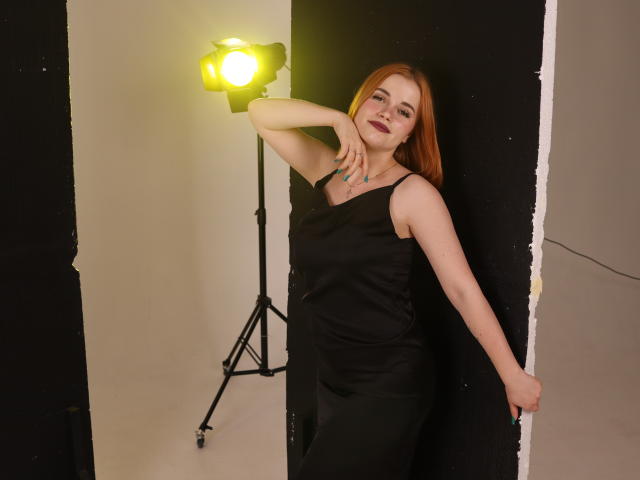 MargaritaKis with red hair, wearing a black dress, leaning against a wall, hand on head, under yellow studio light, smiling, showing her tits slightly through the dress.