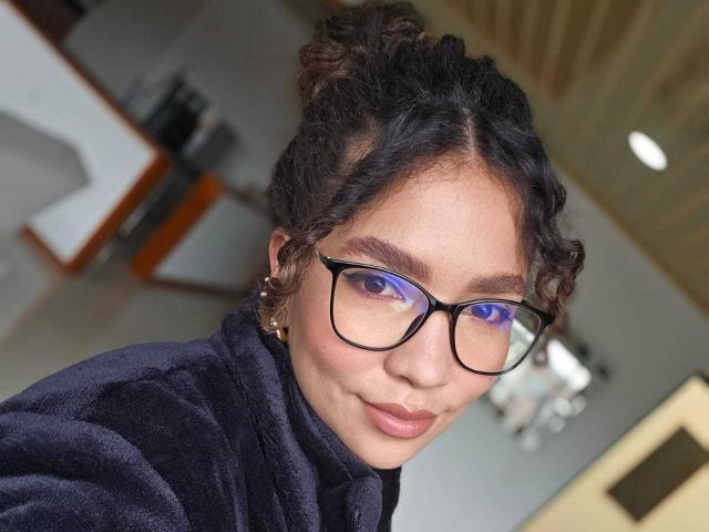 KendallPiierce69 with curly dark hair in buns, wearing black glasses, and a dark purple fleece, smiling at the camera, showing a cute face and a small nose.