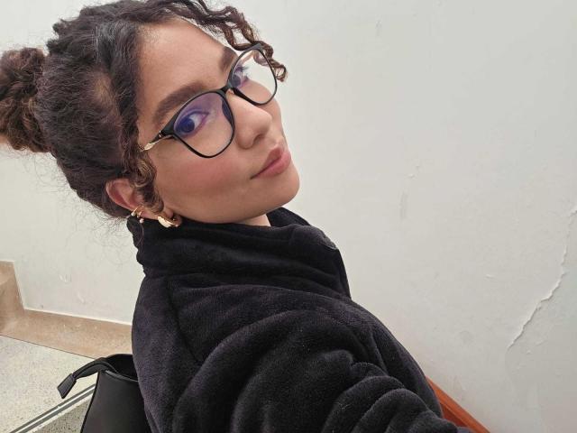 KendallPiierce69 with curly hair in buns, wearing glasses, purple eyeshadow, black hoodie, gold earrings, and a subtle smile, sitting against a white wall.