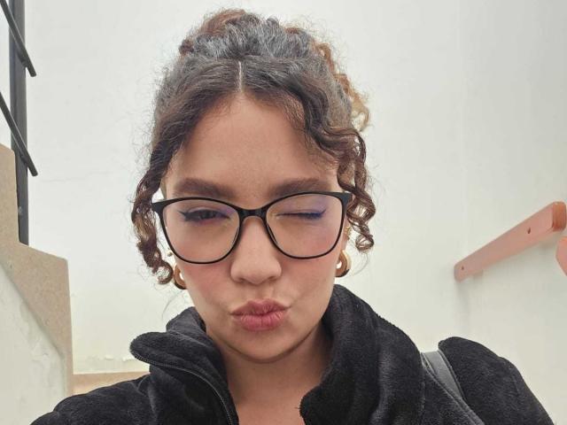 KendallPiierce69 with curly brown hair, black-framed glasses, gold hoop earrings, black jacket, puffy pink lips, and a black backpack, winking. 