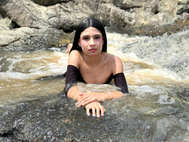 NahimaraSexy, with long black hair, is lying in rocky water, topless with black arm sleeves, her hands in front, water splashing, and rocks around. Boobs, wet, serious expression.
