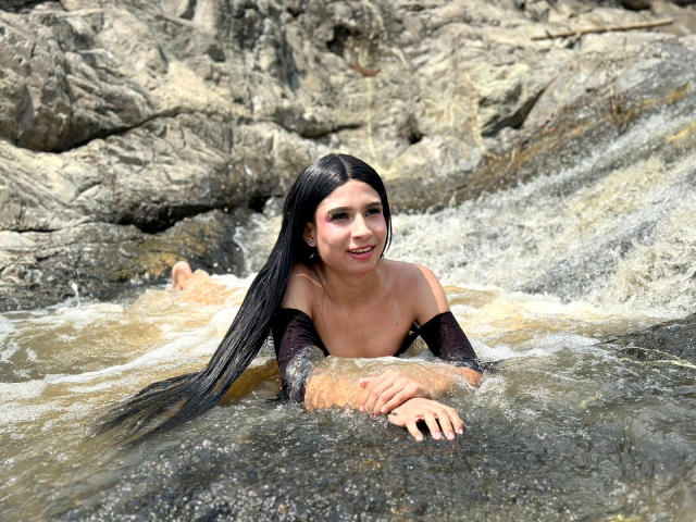 NahimaraSexy with long black hair, topless, in rocky stream, smiling, water splashing, arms resting on rocks, wet skin, visible nipples, water flowing around her.