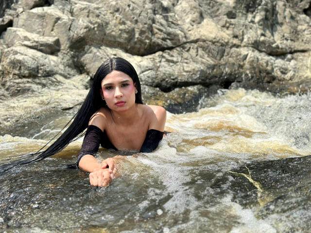 NahimaraSexy with long black hair, pink eye makeup, and bare shoulders, floating in rocky water, arms in front, looking at camera, wearing black off-shoulder top, water splashing.