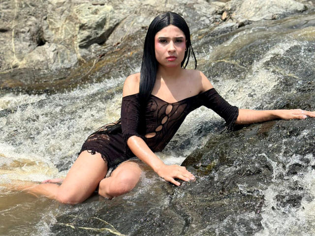 NahimaraSexy with black hair, wearing a see-through black dress, sitting in a rocky stream, legs in water, arms resting on rocks, water splashing around her, looking serious.