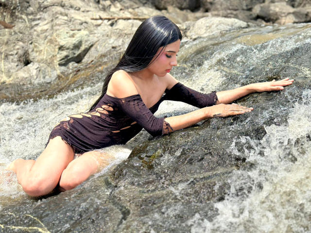 NahimaraSexy, with long black hair, is on her knees in a rocky stream, wearing a wet, black, off-shoulder, cut-out dress, reaching forward with her hands.