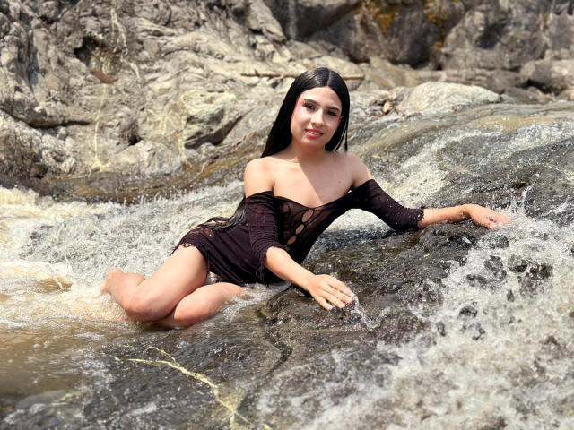 NahimaraSexy with long black hair, sitting in a rocky stream, wearing a wet, black, off-shoulder dress, exposing her small tits and bare ass, with water flowing around her.