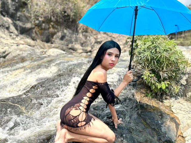 NahimaraSexy, with long black hair, kneels on rocky shore, wearing a black, cut-out one-piece, holding a blue umbrella, looking back with pouty lips, wet skin, and a sexy pose.