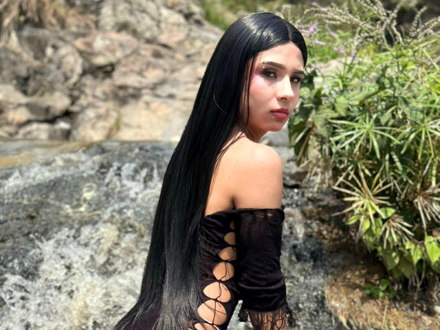 NahimaraSexy with long black hair, off-shoulder black lace-up dress, looking back, outdoors, rocks, green plant, serious expression, pale skin, small nose, dark eyes, slim build, sexy.