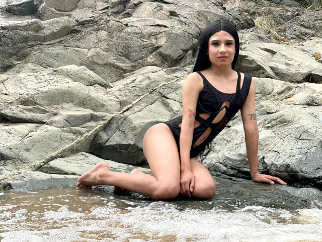 NahimaraSexy with long black hair, cut-out black swimsuit, small tattoos, kneeling on rocky beach, water splashing, bare feet, sexy, pale skin, cute, slim, small breasts.
