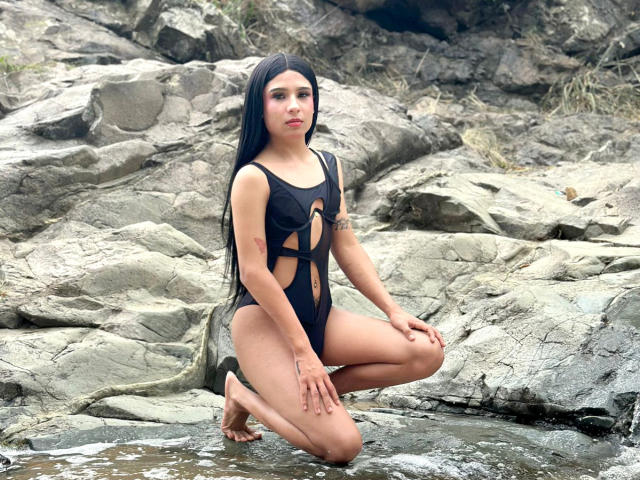 NahimaraSexy with long black hair, kneeling on rocks in a black cut-out swimsuit, showing her small tits and narrow ass. Water is around her knees. She's looking at the camera.